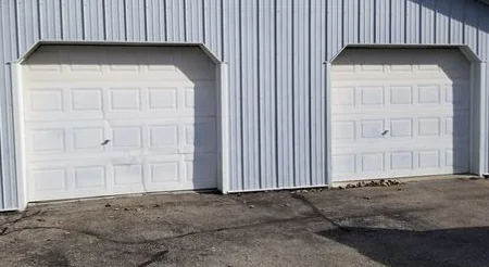 Two white raised-panel garage doors without windows in a light gray steel-sided pole building, above a cracked asphalt apron.