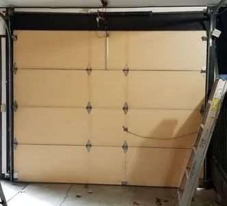 Inside view of a tan garage door with hinges and a loose cable across it, with a ladder leaning at the right.