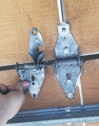 A hand points to a bent garage door hinge next to an intact hinge on a wood-grain door section.