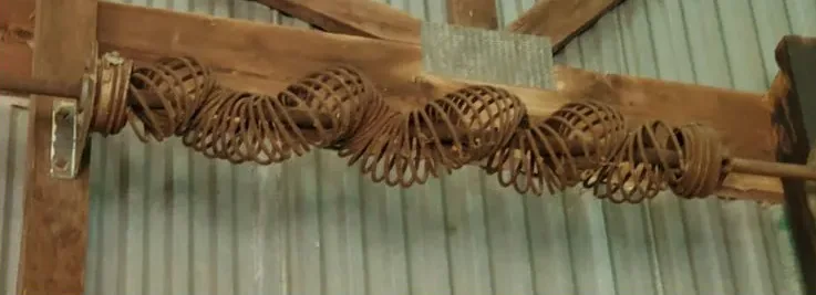 A rusted, stretched coil spring running along a wooden beam against corrugated metal siding.
