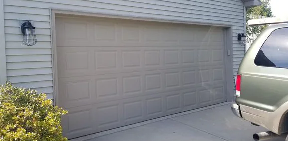 A plain tan double garage door with raised panels on a gray-sided house, with a parked SUV at the right edge.