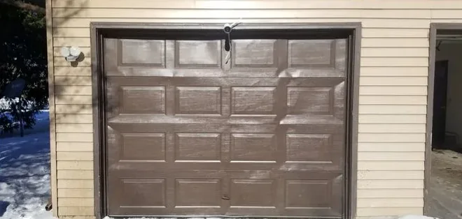 A brown raised-panel garage door in beige siding, with snow on the ground.