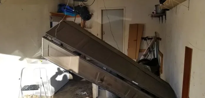 Inside a cluttered garage, a brown sectional door hangs out of its opening at a steep angle.