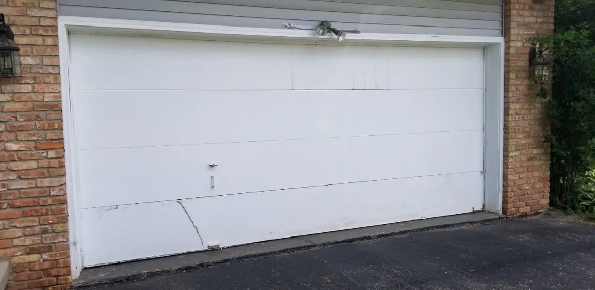 A white double garage door between brick walls and coach lights, with a repaired crack still visible in the bottom section.