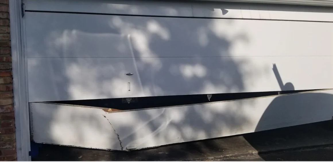 A white garage door in dappled shade beside a brick wall, its bottom section bent outward and split open.