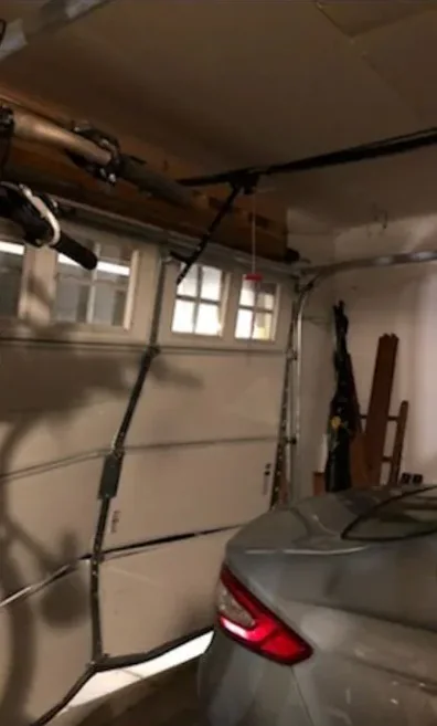 Inside view of a garage door with a row of windows and bent sections, behind hanging bicycles, with a parked car at the right.